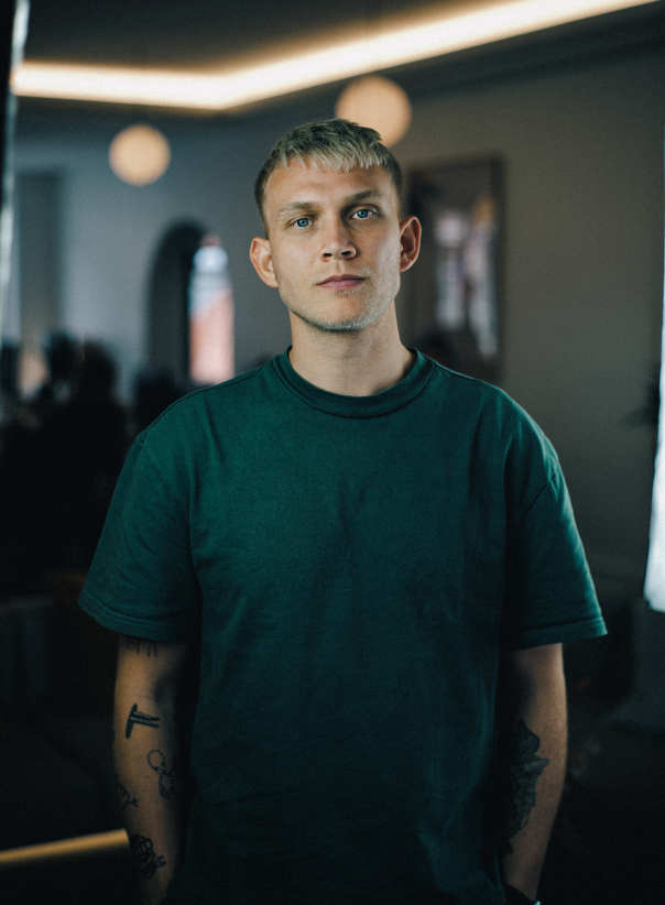 Young man with blonde hair, blue eyes, and tattoos on his arms wearing a dark green t-shirt in a dimly lit room.